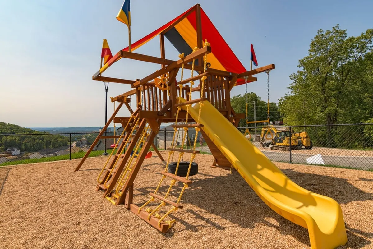 A bright and cheerful outdoor playground featuring a colorful structure with a yellow slide and vibrant flags, surrounded by open space for play and family gatherings. Children can be seen delighting in the fun environment.