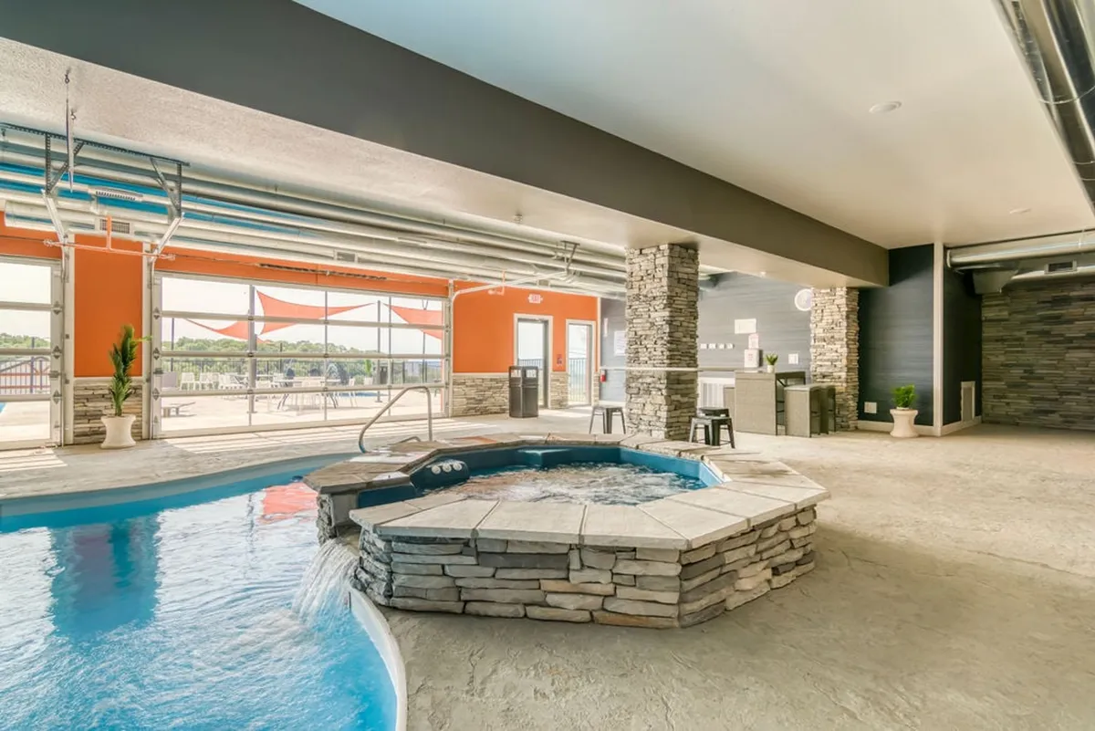 This interior image features a luxurious hot tub and pool area with expansive views through large windows. The setting includes a decorative stone wall, bar stools, and a tranquil, social atmosphere—ideal for relaxing after a day of activities. The windows overlook a scenic landscape, blending the interior comforts with the beauty of the outdoors.
