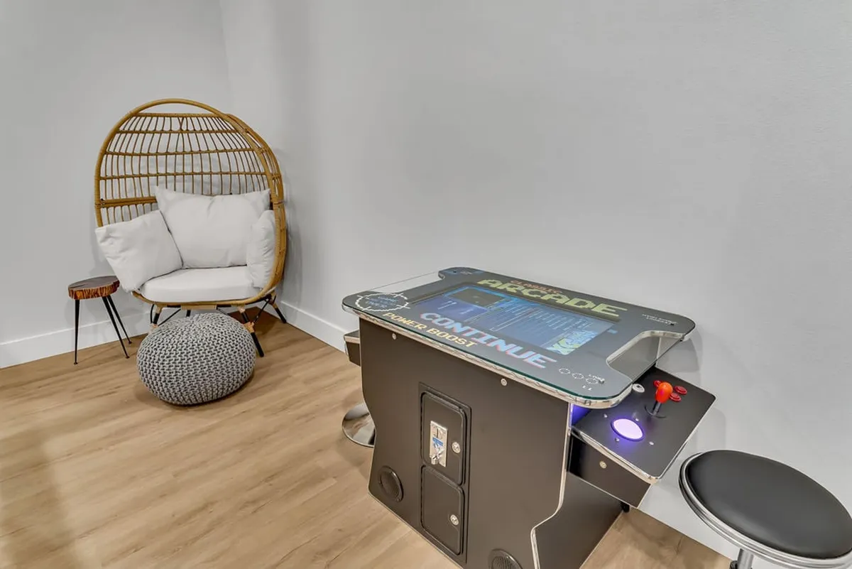 A welcoming interior corner featuring a retro-style cocktail arcade game, a rattan chair, and a pouf, set against a neutral-toned wall. The space radiates playful charm and encourages both solo and social entertainment.
