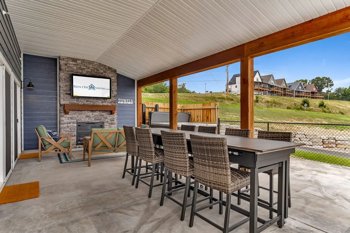 An exterior shot of a stylish outdoor dining area centered around a stone fireplace with a mounted TV. The setup encourages social gatherings, outdoor meals, and cozy evenings by the fire. Ample seating and family-friendly space invite guests to linger, while the fireplace serves as both a focal point and a source of warmth for year-round enjoyment.