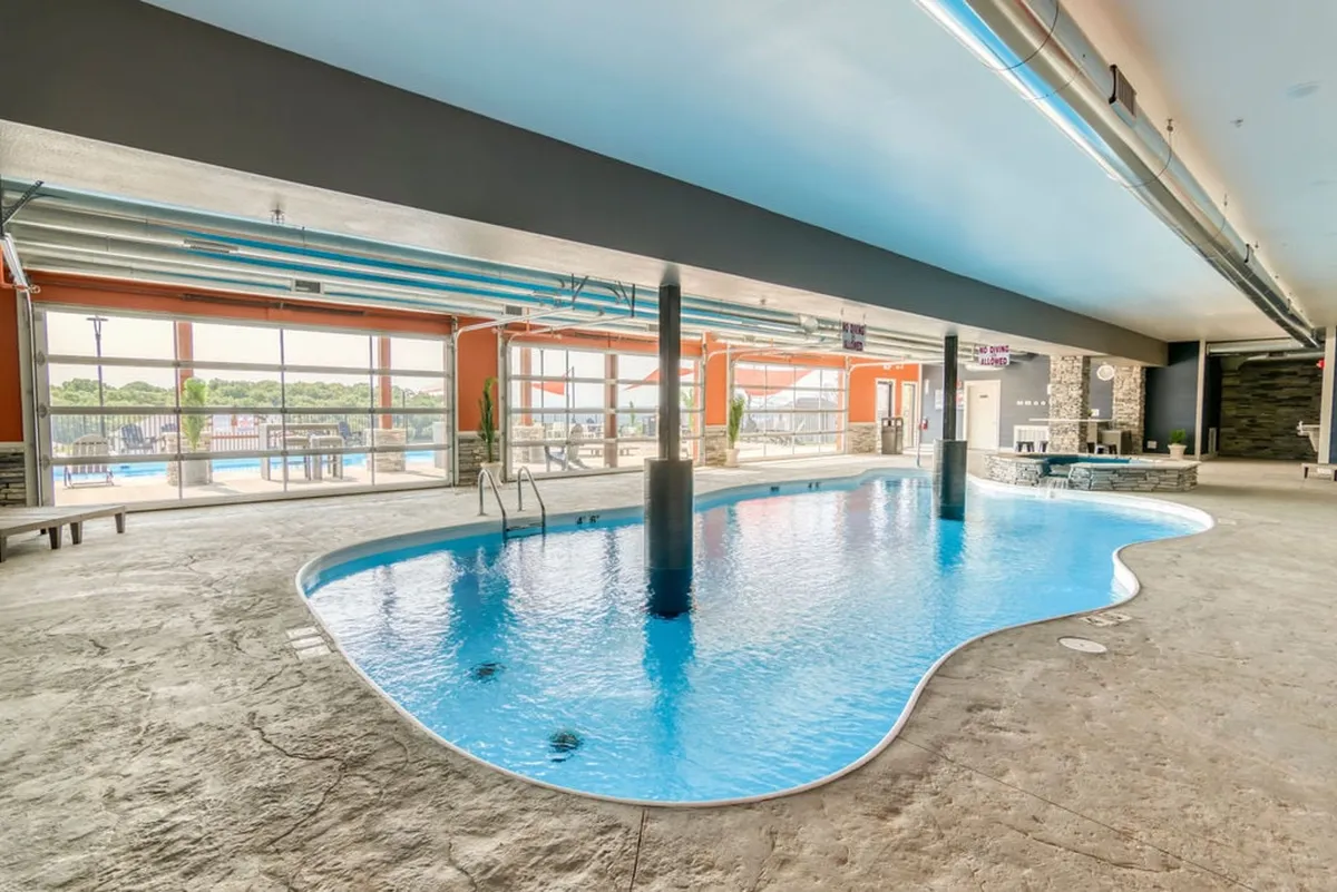 A spacious indoor pool area featuring modern lighting, large windows that let in natural light, a tranquil pool surrounded by a stone wall, and a cozy seating area. The atmosphere is serene and rejuvenating, suitable for year-round swimming and relaxation.