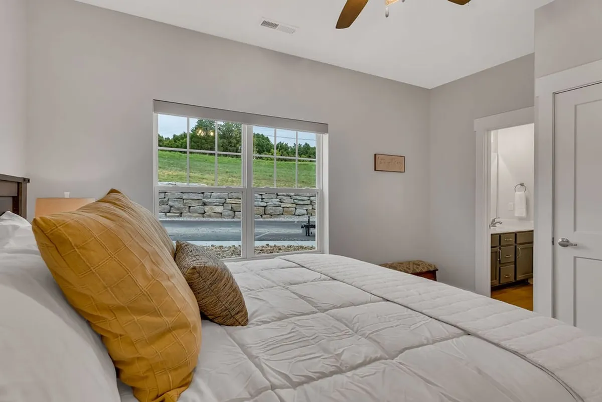 A serene bedroom with a plush bed adorned with decorative pillows, a large window filling the space with natural light, and an entrance to a private bathroom. The soothing palette and soft textures create an inviting atmosphere perfect for relaxation.