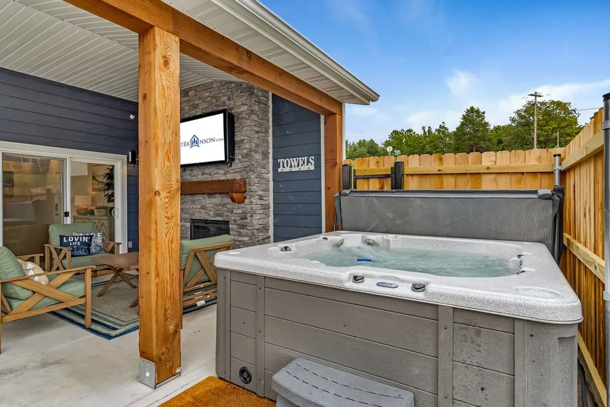 This exterior image spotlights a tranquil patio setup featuring a bubbling hot tub, a modern outdoor seating area, and a prominent fireplace below a mounted TV. The surrounding elements, such as soft outdoor lighting and natural decor, evoke a sense of comfort and relaxation while still being clearly oriented outdoors. The lake is visible in the background, reinforcing the sought-after lakeview and enhancing the appeal of the outdoor amenities.